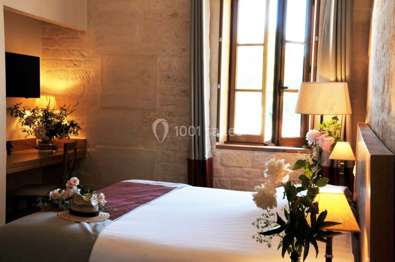 Chambre lumineuse avec lit double, bureau, lampe et fenêtre en bois, décorée de fleurs et éclairée par une lumière naturelle.