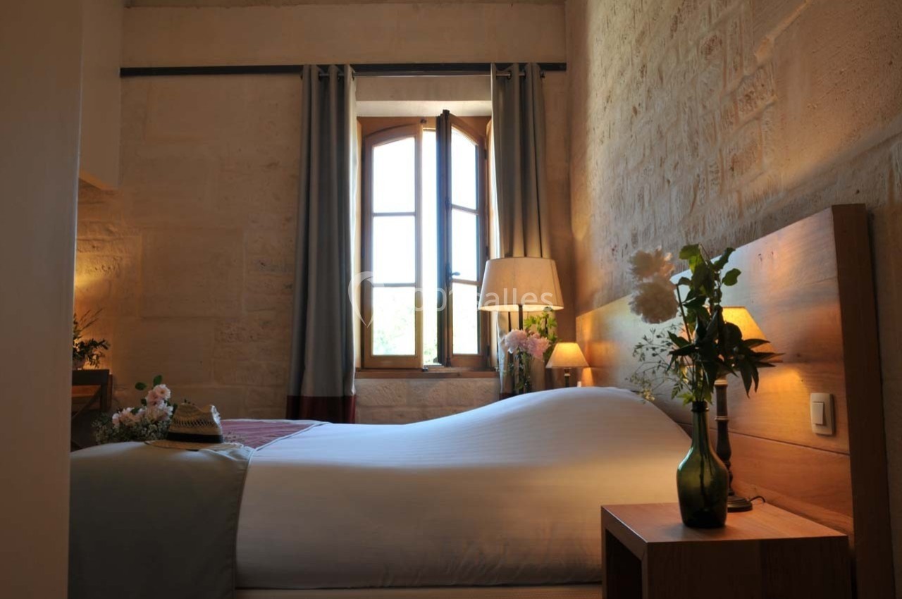 Chambre lumineuse avec lit double, mur en pierre, fenêtre ouverte, rideaux, lampe de chevet et vase de fleurs.