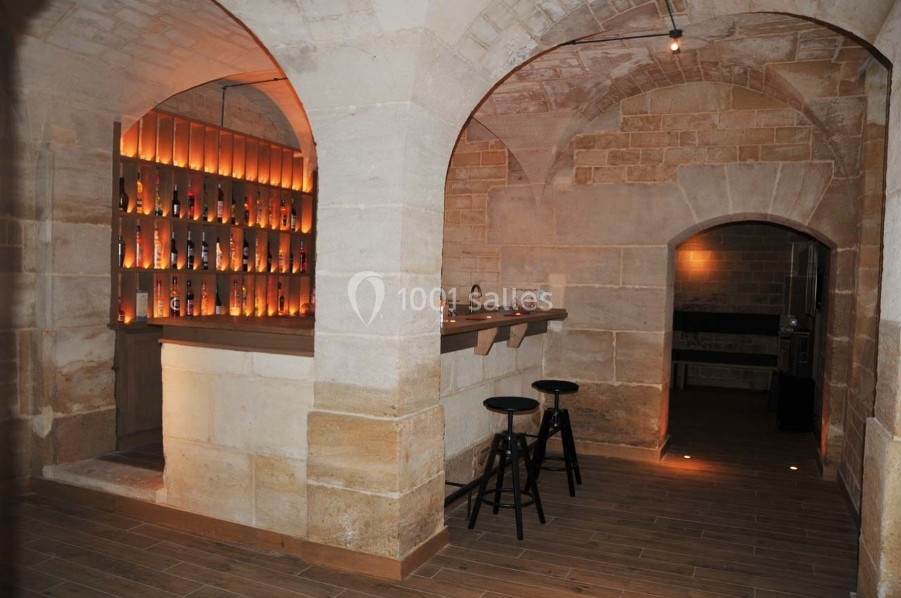 Cave voûtée en pierre avec un bar éclairé, des tabourets noirs et des étagères remplies de bouteilles.