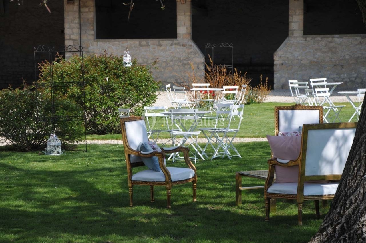 Chaises et fauteuils disposés sur une pelouse, entourés de buissons et de tables blanches dans un jardin extérieur.