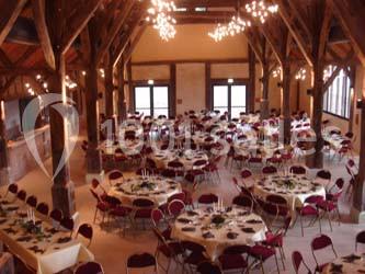 Salle de réception avec charpente en bois, tables rondes dressées et chaises rouges, éclairée par des lustres suspendus.