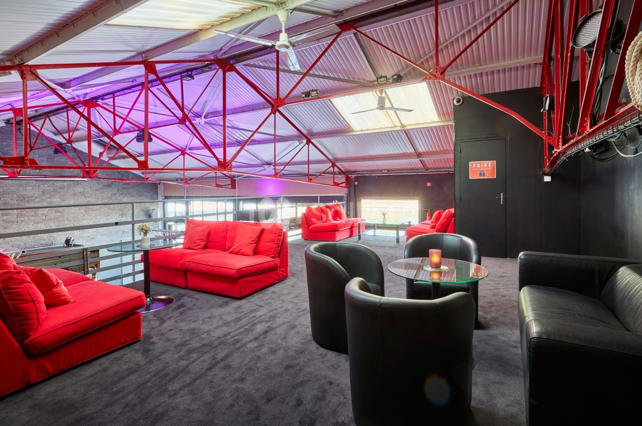 Espace lounge avec canapés rouges et noirs, tables basses et structure métallique apparente sous un toit en pente.