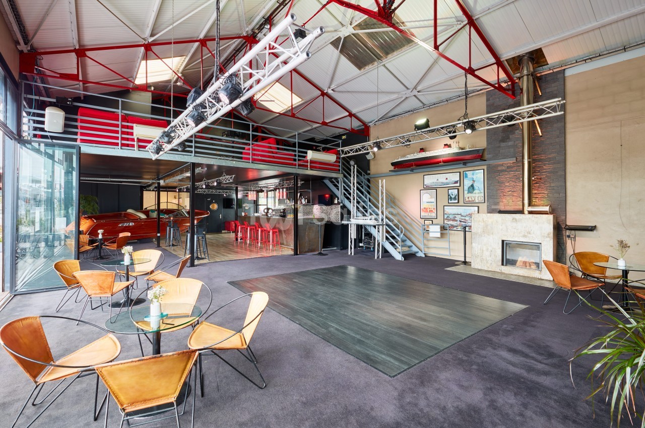 Salle spacieuse avec mezzanine, tables en verre, chaises design, bar rouge, cheminée et grande baie vitrée.