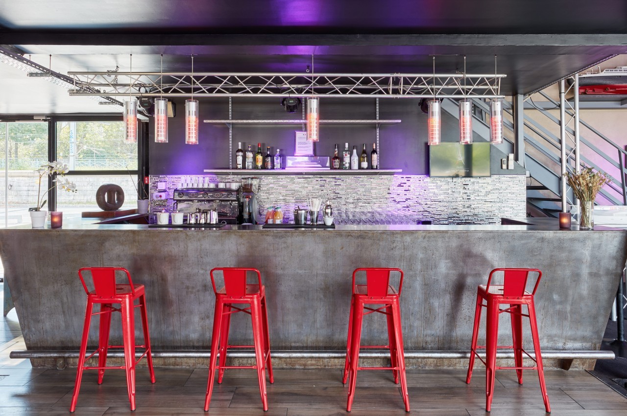 Bar moderne avec comptoir en métal, tabourets rouges, éclairage violet et étagères garnies de bouteilles.