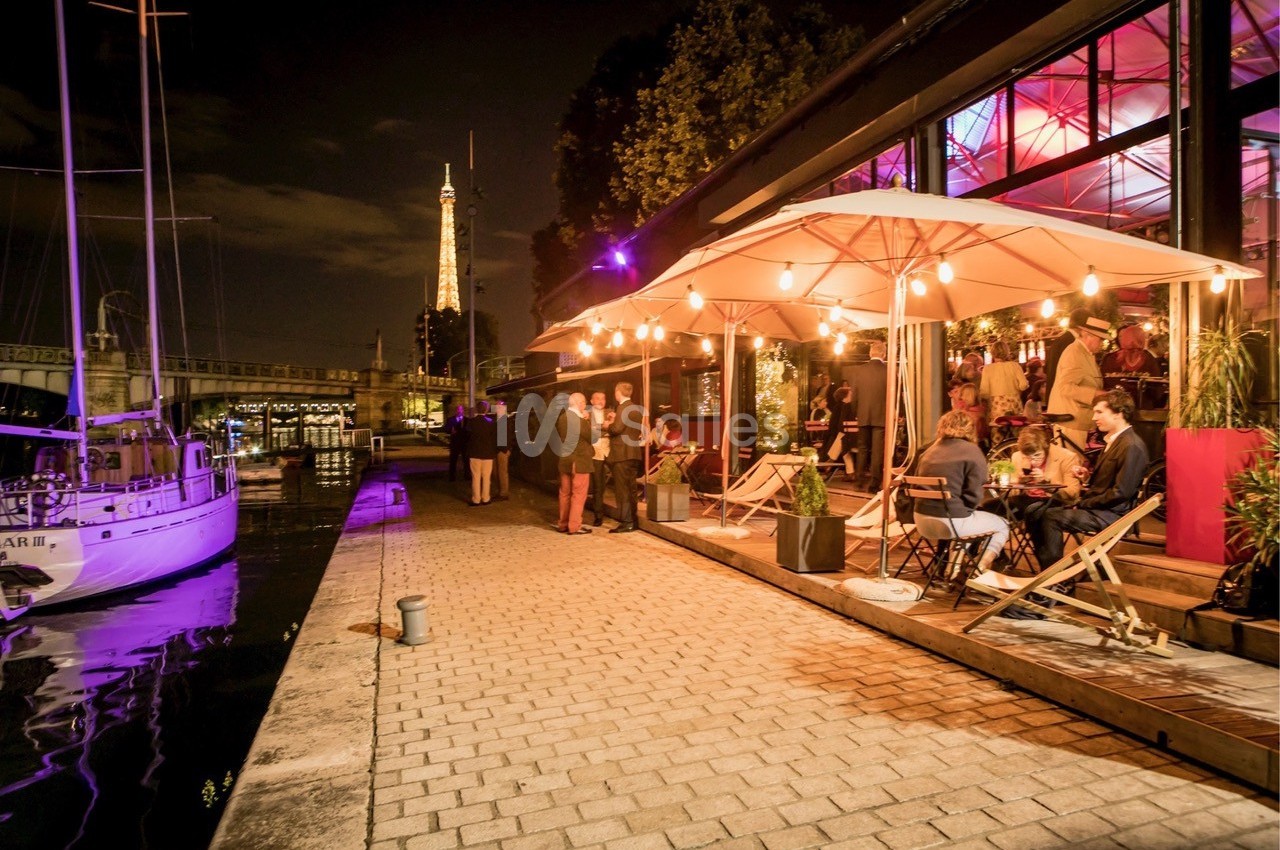 Terrasse animée d'un restaurant au bord de l'eau, éclairée par des guirlandes lumineuses, avec la Tour Eiffel en arrière…