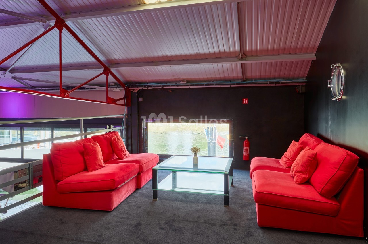 Deux canapés rouges autour d'une table basse en verre dans un espace intérieur avec vue sur l'extérieur par une fenêtre.