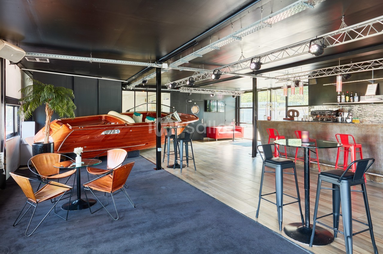 Salle moderne avec bar, tables hautes, fauteuils design et un bateau en bois intégré dans la décoration.