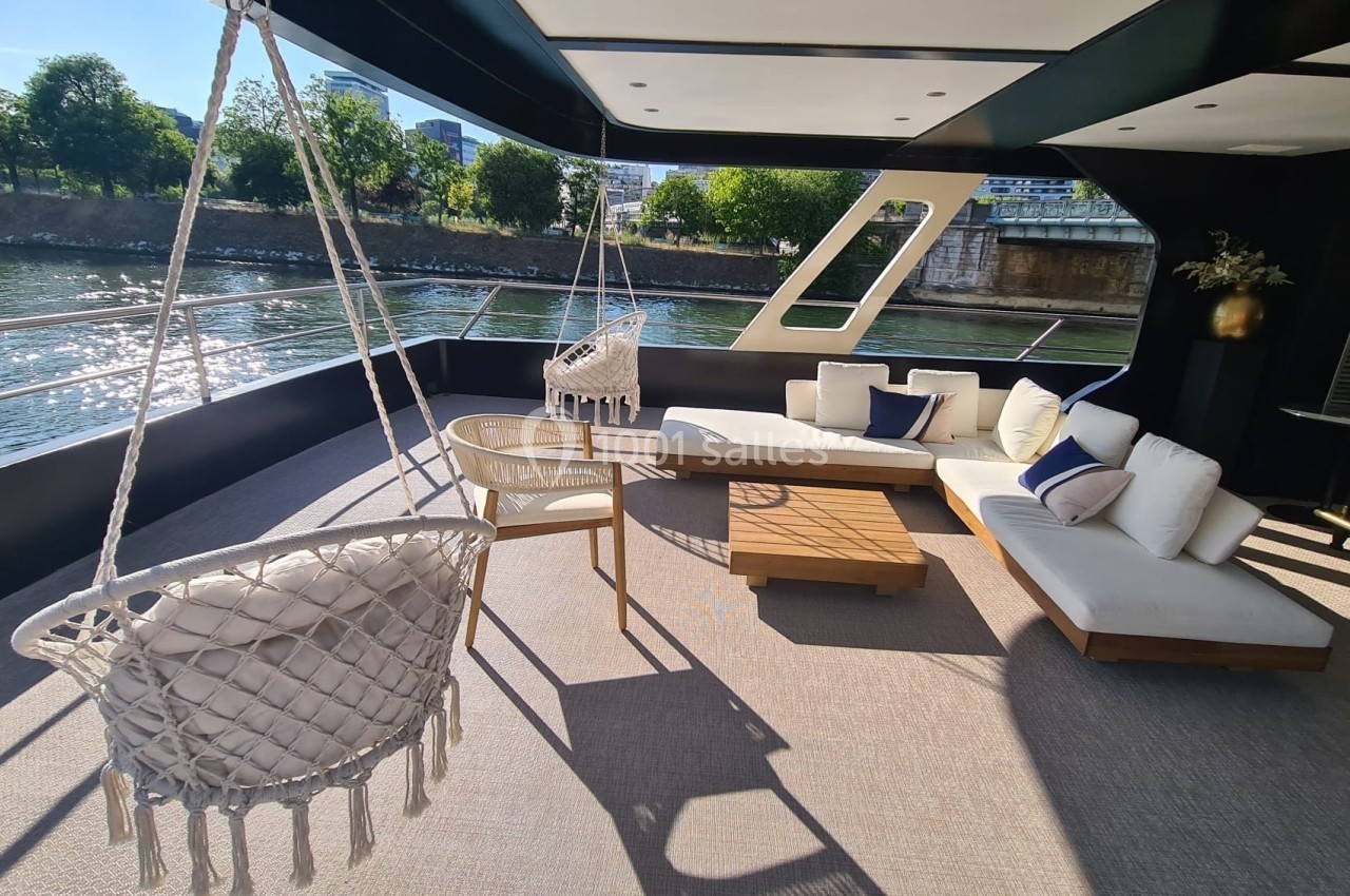 Espace extérieur d'un bateau avec fauteuils suspendus, canapé blanc, table basse en bois et vue sur une rivière bordée d…