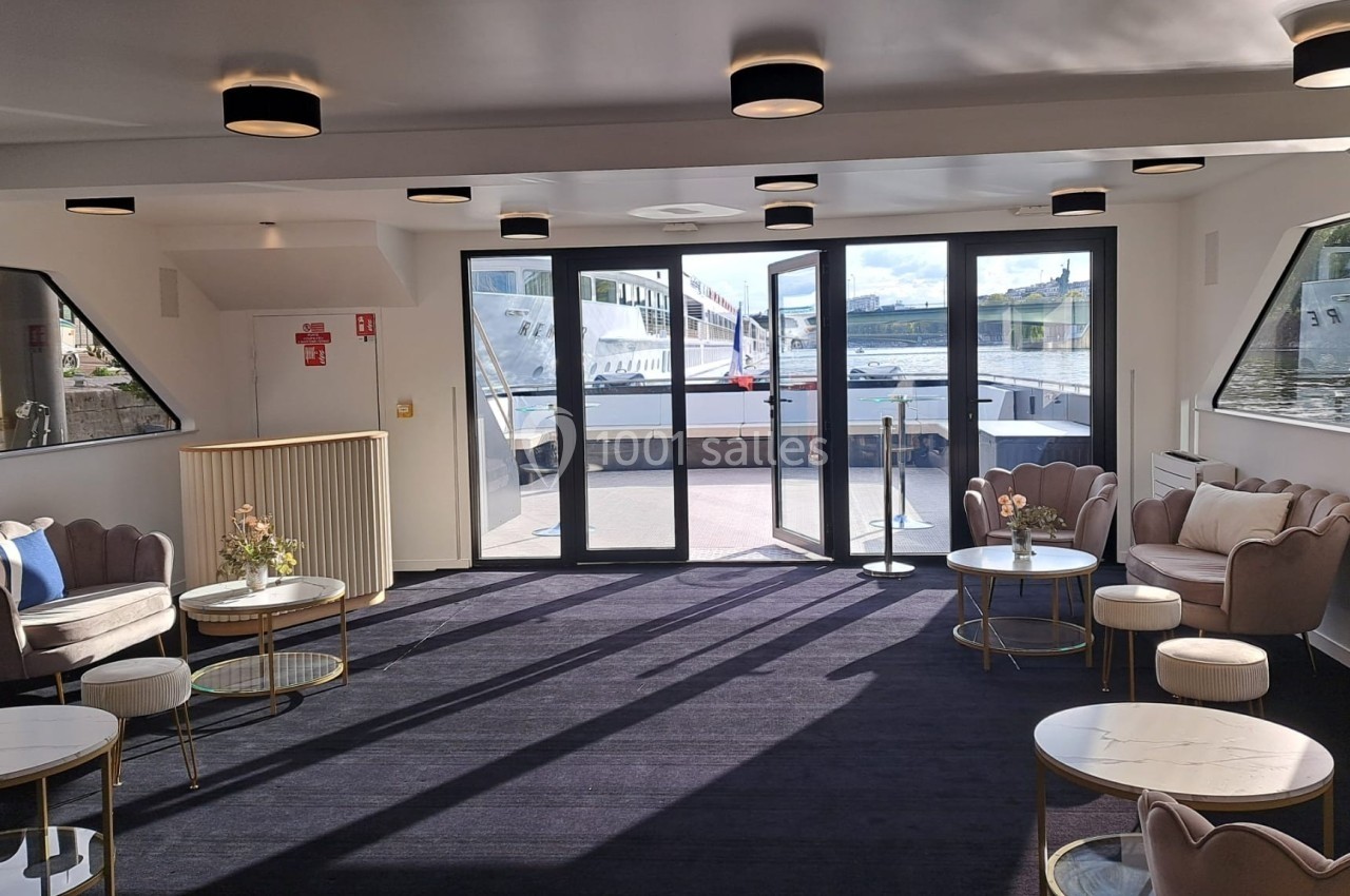 Salon lumineux avec fauteuils, tables basses et grandes baies vitrées donnant sur un quai et un bateau amarré.