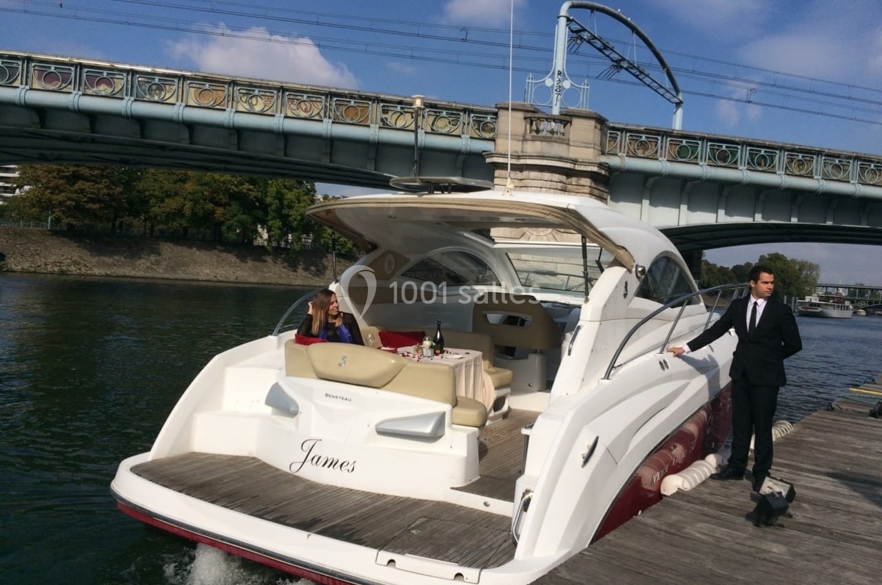 Un bateau de plaisance amarré à un quai, avec une femme assise à bord et un homme en costume debout sur le ponton.