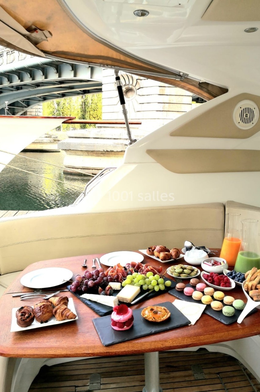 Table garnie de viennoiseries, fruits, macarons et jus, installée sur un bateau près d'un canal urbain.