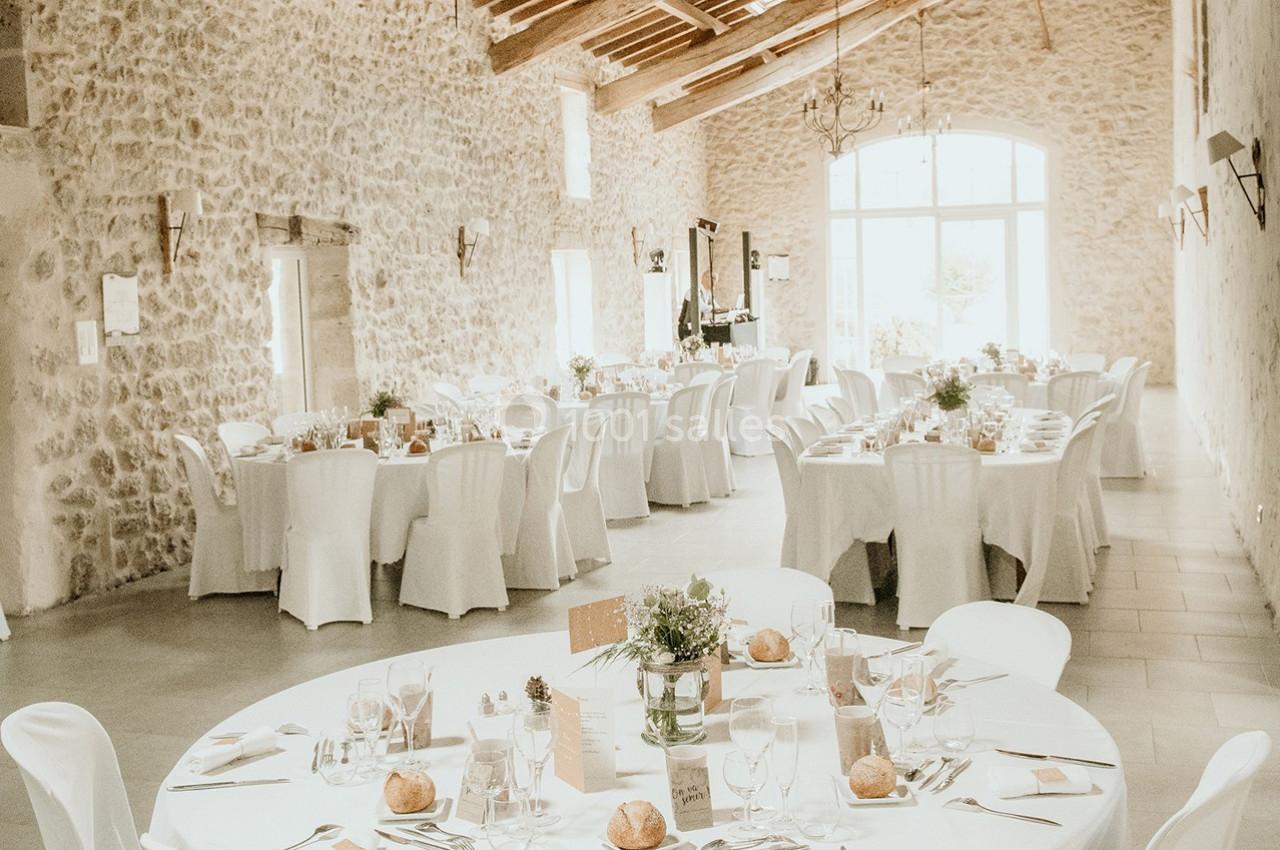Salle de réception lumineuse avec des tables rondes dressées, nappes blanches et décoration florale.