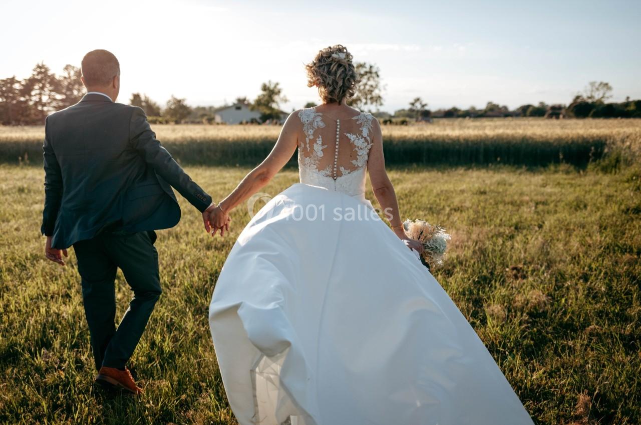 Un couple en tenue de mariage marche main dans la main dans un champ au coucher du soleil.