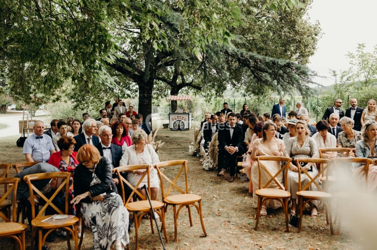 Invités assis en plein air sous des arbres lors d'une cérémonie, avec une décoration champêtre en arrière-plan.