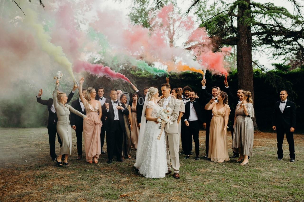 Un couple de mariés entouré d'invités tenant des fumigènes colorés dans un jardin.
