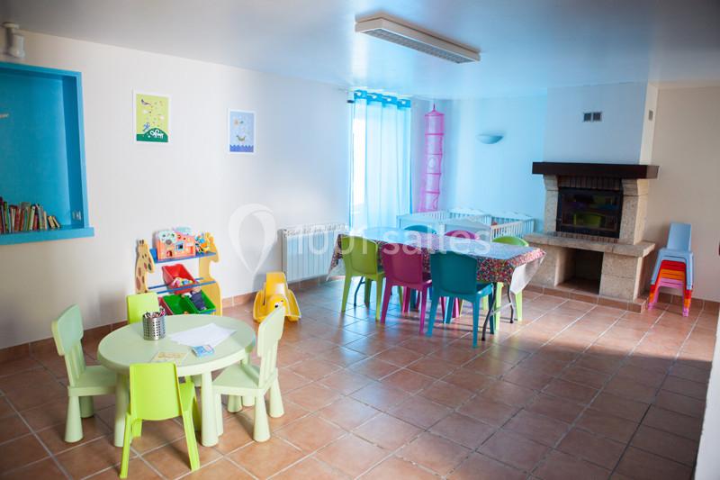 Salle lumineuse avec tables et chaises colorées pour enfants, étagères, cheminée et sol en carrelage.