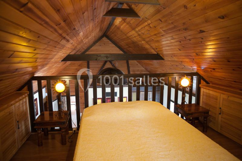 Chambre mansardée avec plafond en bois, lit couvert d'une couette jaune et deux lampes de chevet allumées.