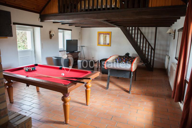 Salle de jeux avec table de billard, baby-foot, canapé, télévision et escalier en bois menant à une mezzanine.