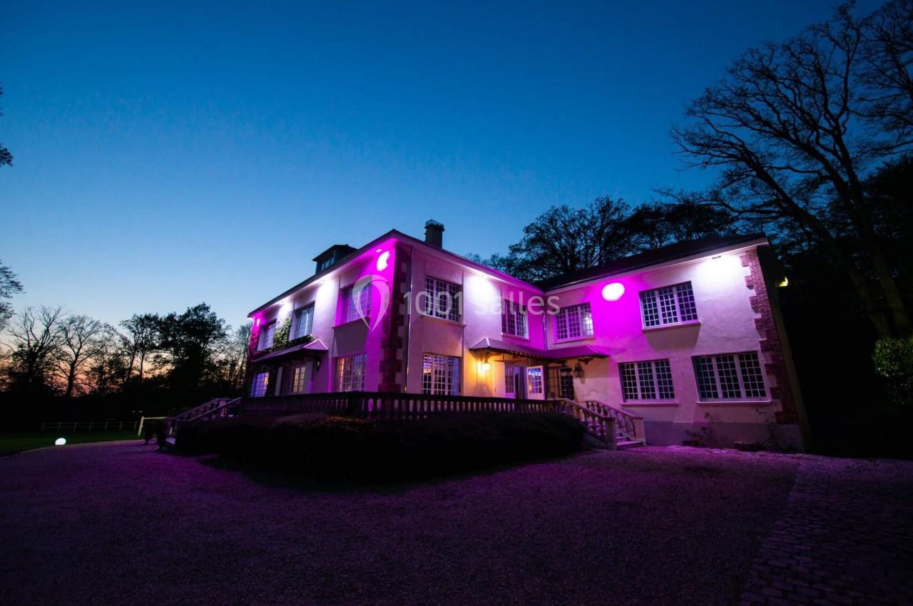 Façade d'une maison éclairée par des lumières roses et violettes, entourée d'arbres au crépuscule.