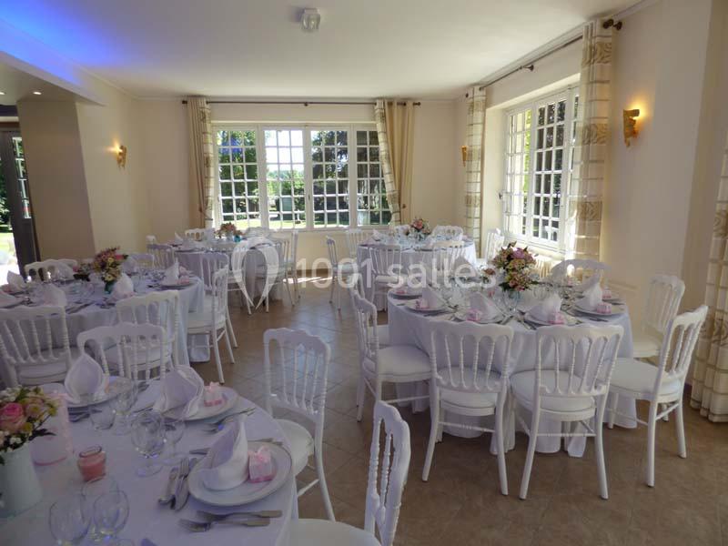 Salle lumineuse décorée pour un événement, avec tables rondes dressées, nappes blanches et arrangements floraux.