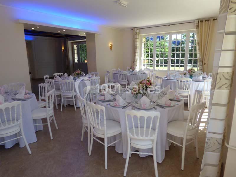 Salle de réception lumineuse avec tables rondes dressées, nappes blanches et décorations florales.