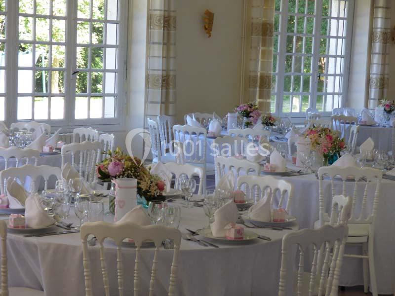 Salle de réception lumineuse avec des tables rondes décorées de nappes blanches, fleurs et serviettes pliées.