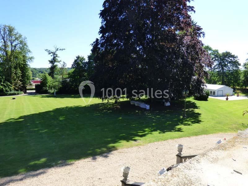 Pelouse verdoyante avec un grand arbre au centre, entourée de végétation et d'un bâtiment blanc en arrière-plan.