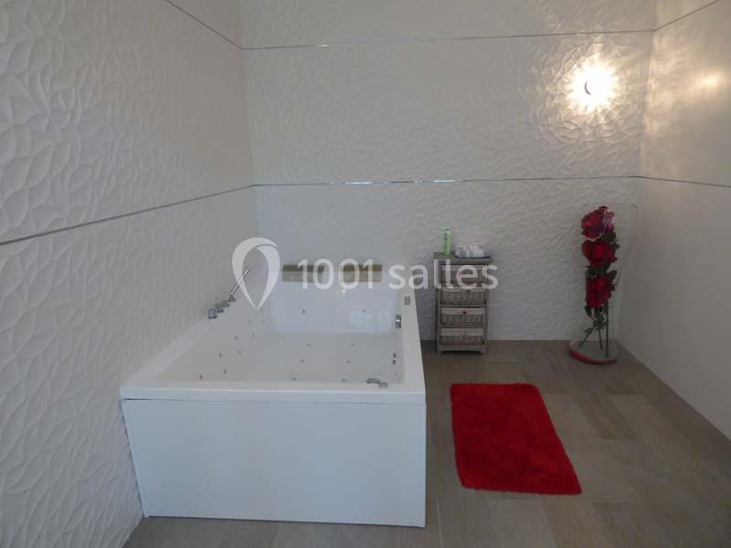 Salle de bain moderne avec une baignoire blanche, un tapis rouge, un mur texturé et un vase de fleurs rouges.