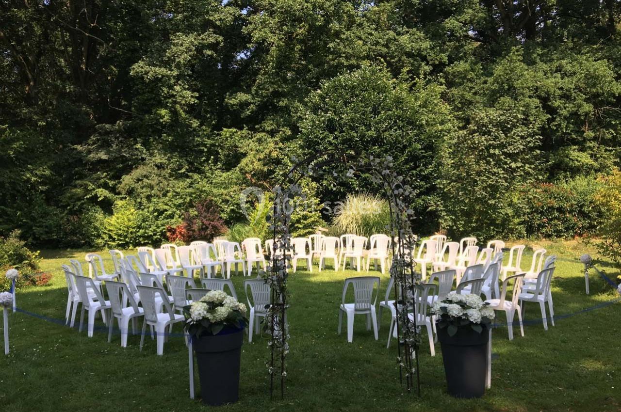 Chaises blanches disposées en cercle autour d'une arche fleurie dans un jardin verdoyant.