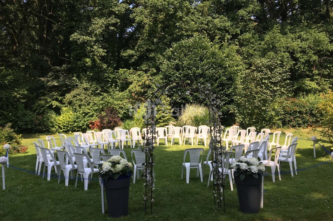 Chaises blanches disposées en cercle autour d'une arche fleurie dans un jardin verdoyant.