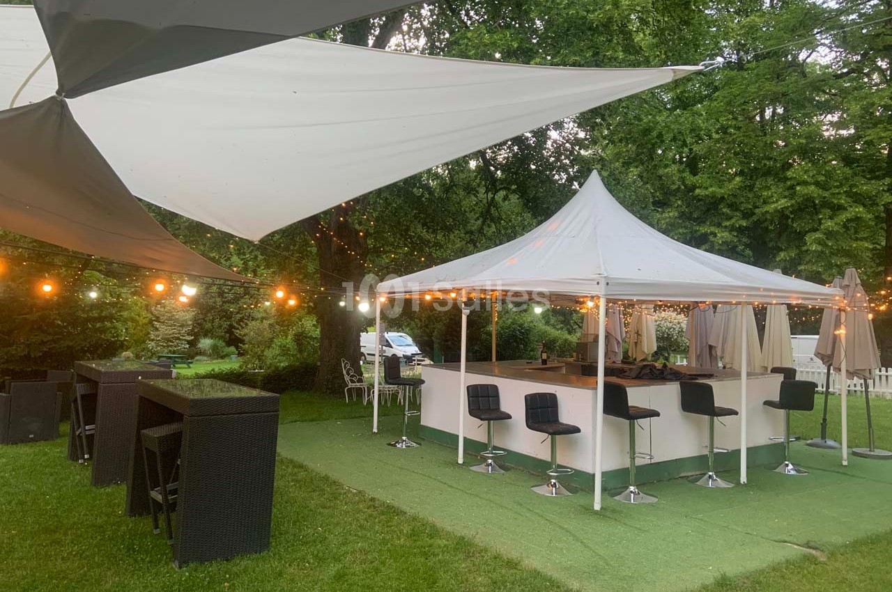 Bar extérieur sous des tentes blanches, entouré de guirlandes lumineuses et de mobilier de jardin sur pelouse.