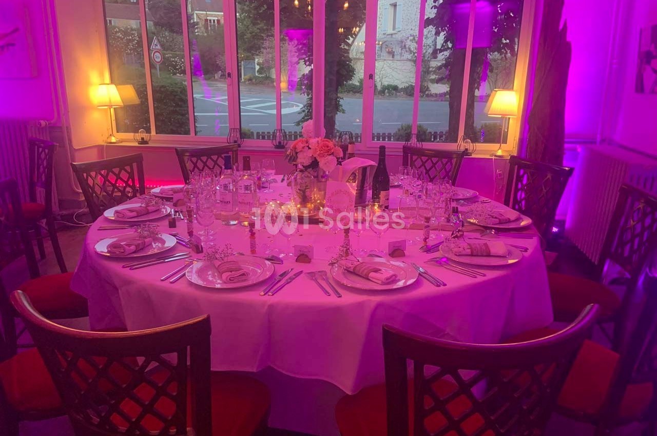 Table ronde élégamment dressée avec nappes blanches, vaisselle raffinée et centre de table floral, éclairée par une lumière…