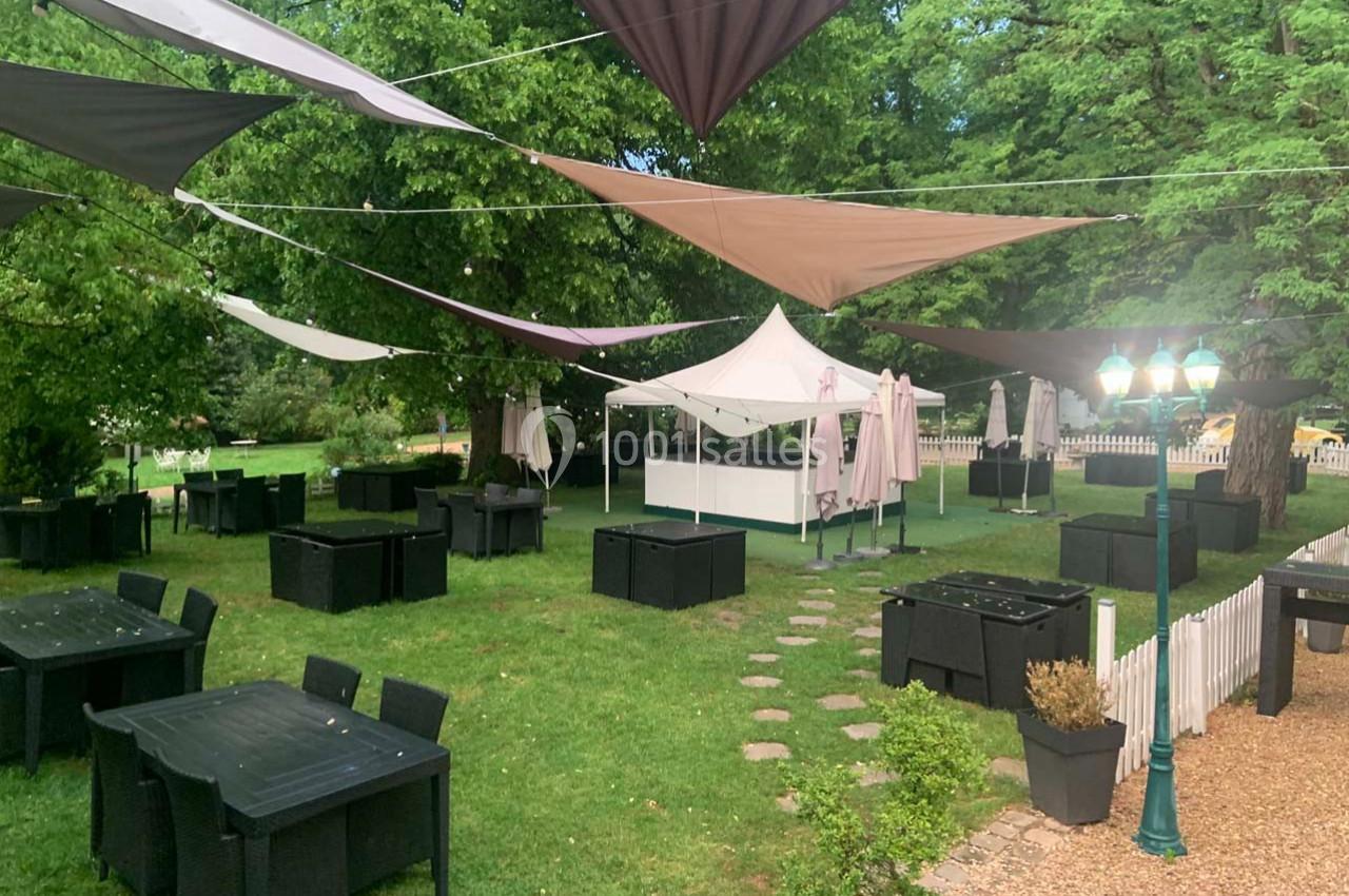 Terrasse extérieure avec tables noires, parasols fermés et voiles d'ombrage, entourée de verdure et éclairée par un…