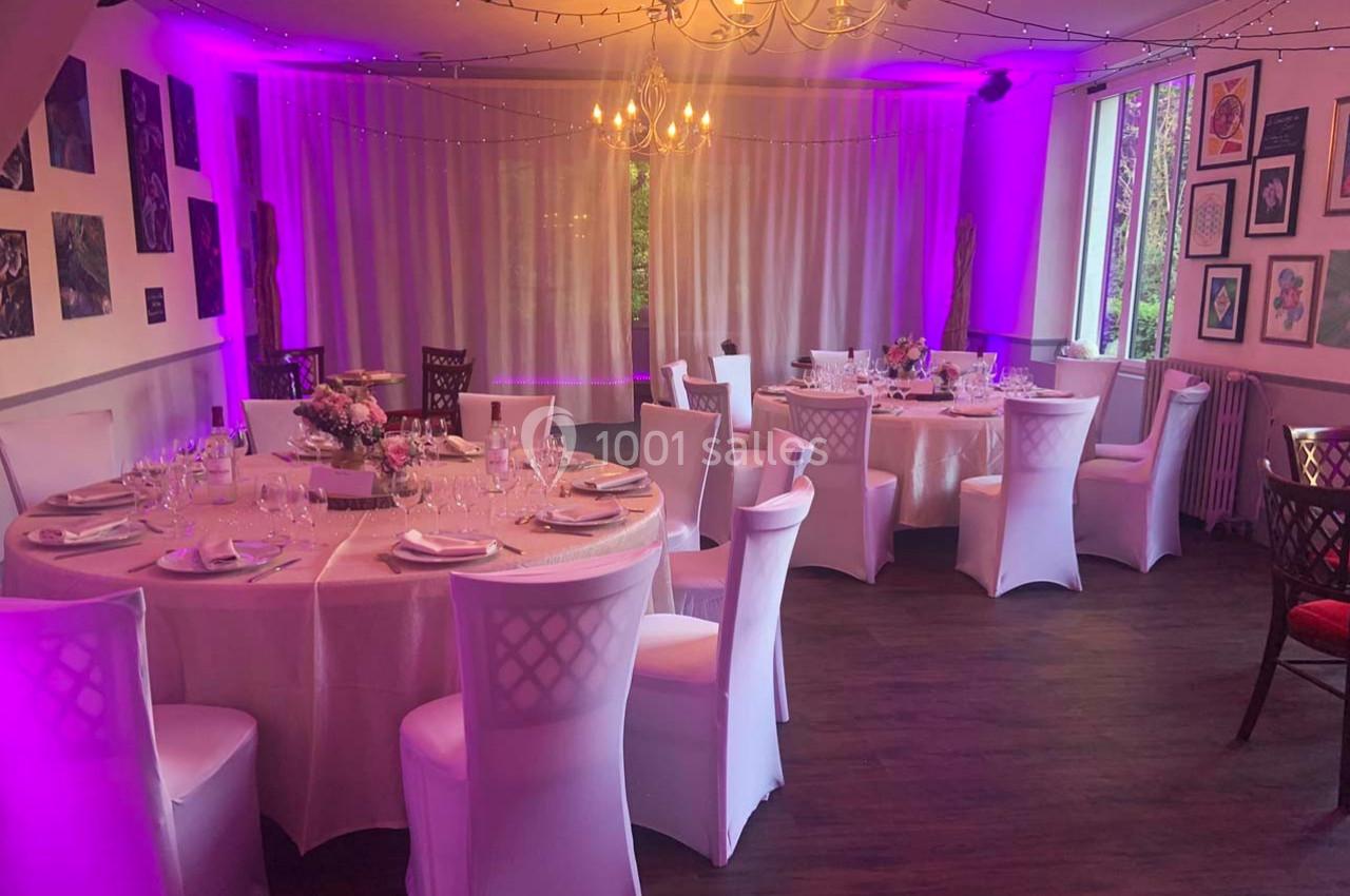 Salle décorée pour un événement, avec tables rondes dressées, chaises blanches et éclairage violet tamisé.