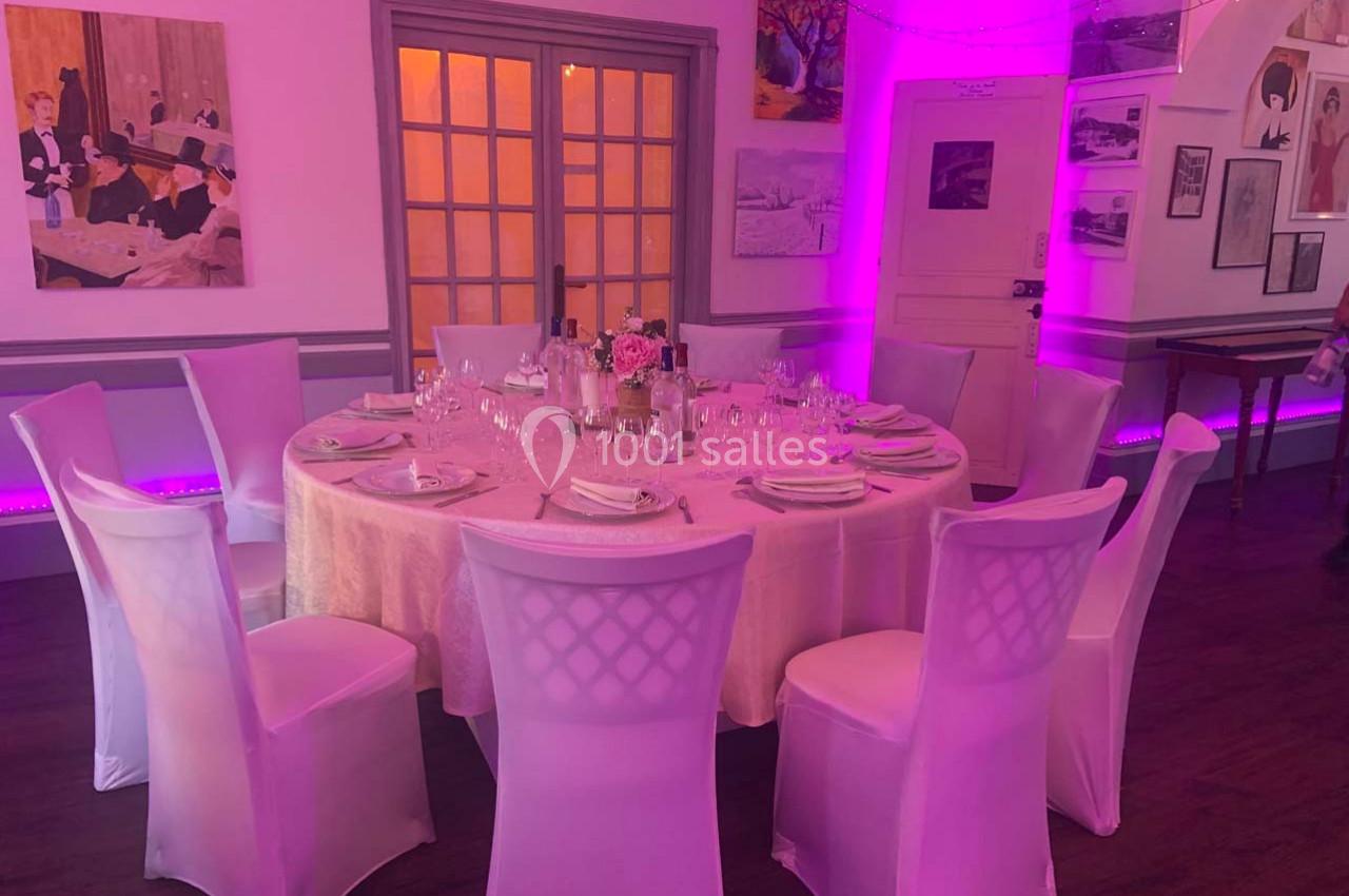 Table ronde dressée avec une nappe rose, entourée de chaises blanches, dans une salle décorée et éclairée en rose.