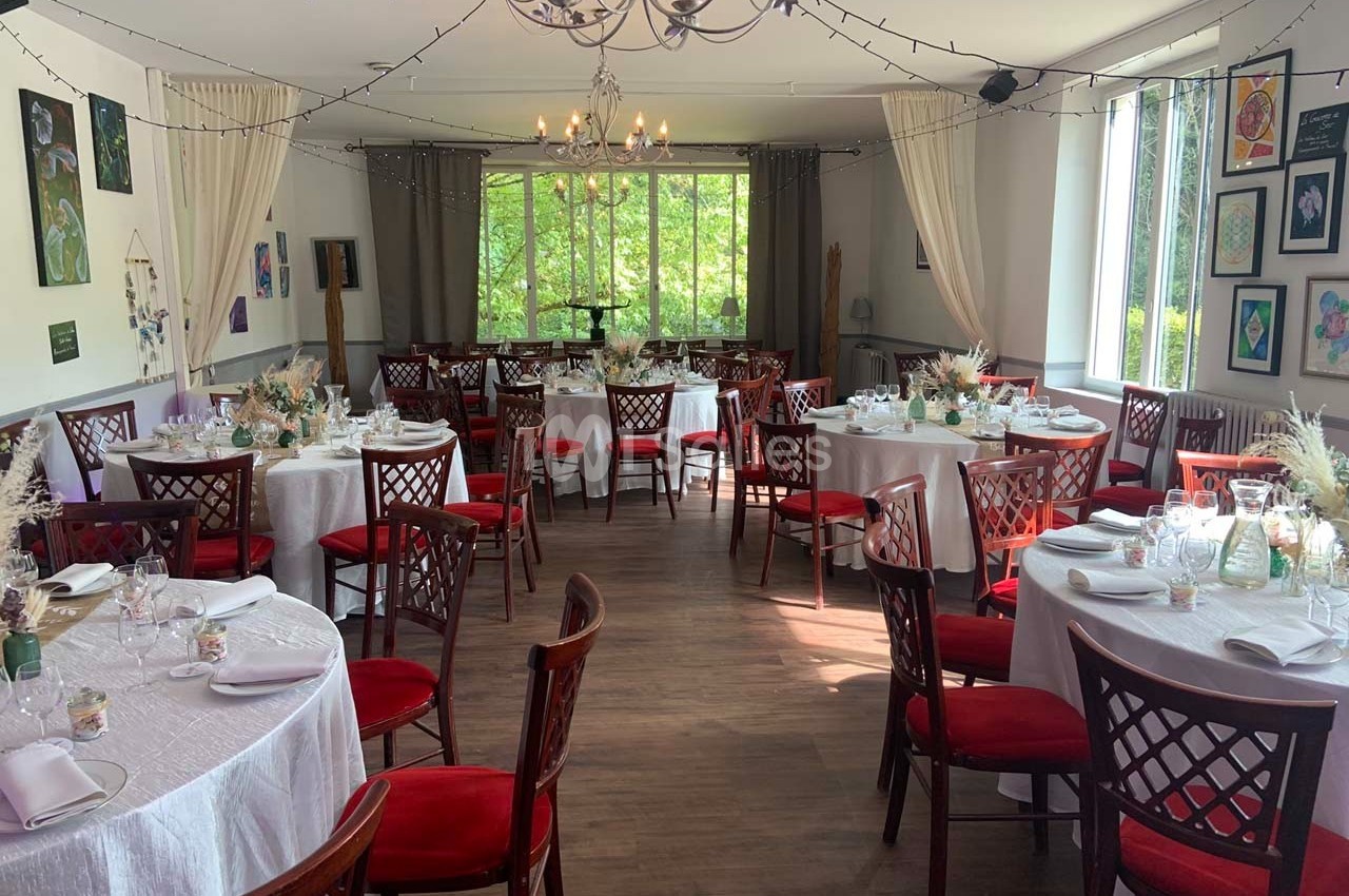Salle de réception lumineuse avec tables rondes dressées, chaises en bois rouge et décorations florales.