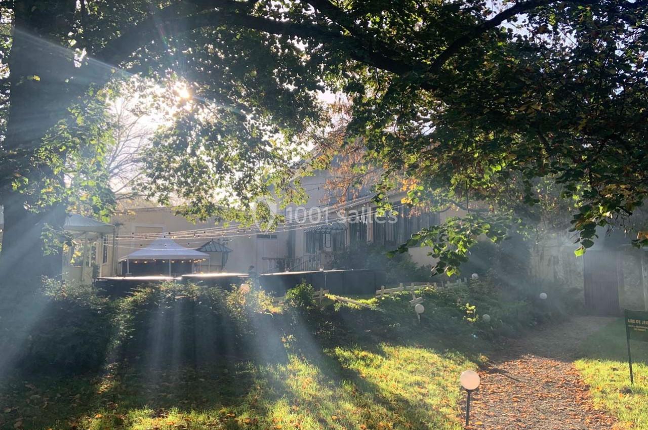 Lumière du soleil traversant les arbres, illuminant un jardin avec des bâtiments en arrière-plan.