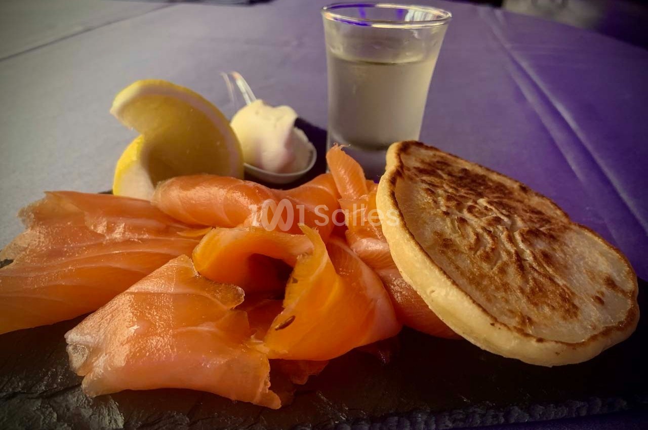 Tranches de saumon fumé avec blini, crème fraîche, citron et un verre de boisson sur une table violette.