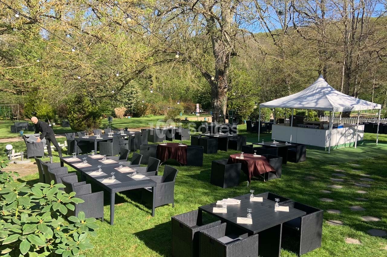 Terrasse extérieure d'un restaurant avec tables noires dressées, située dans un jardin verdoyant sous des arbres.
