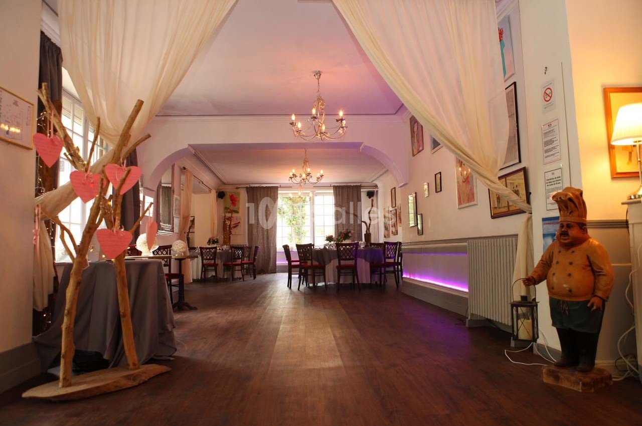 Salle de restaurant lumineuse avec tables dressées, rideaux blancs, décoration en bois et statue de chef cuisinier.