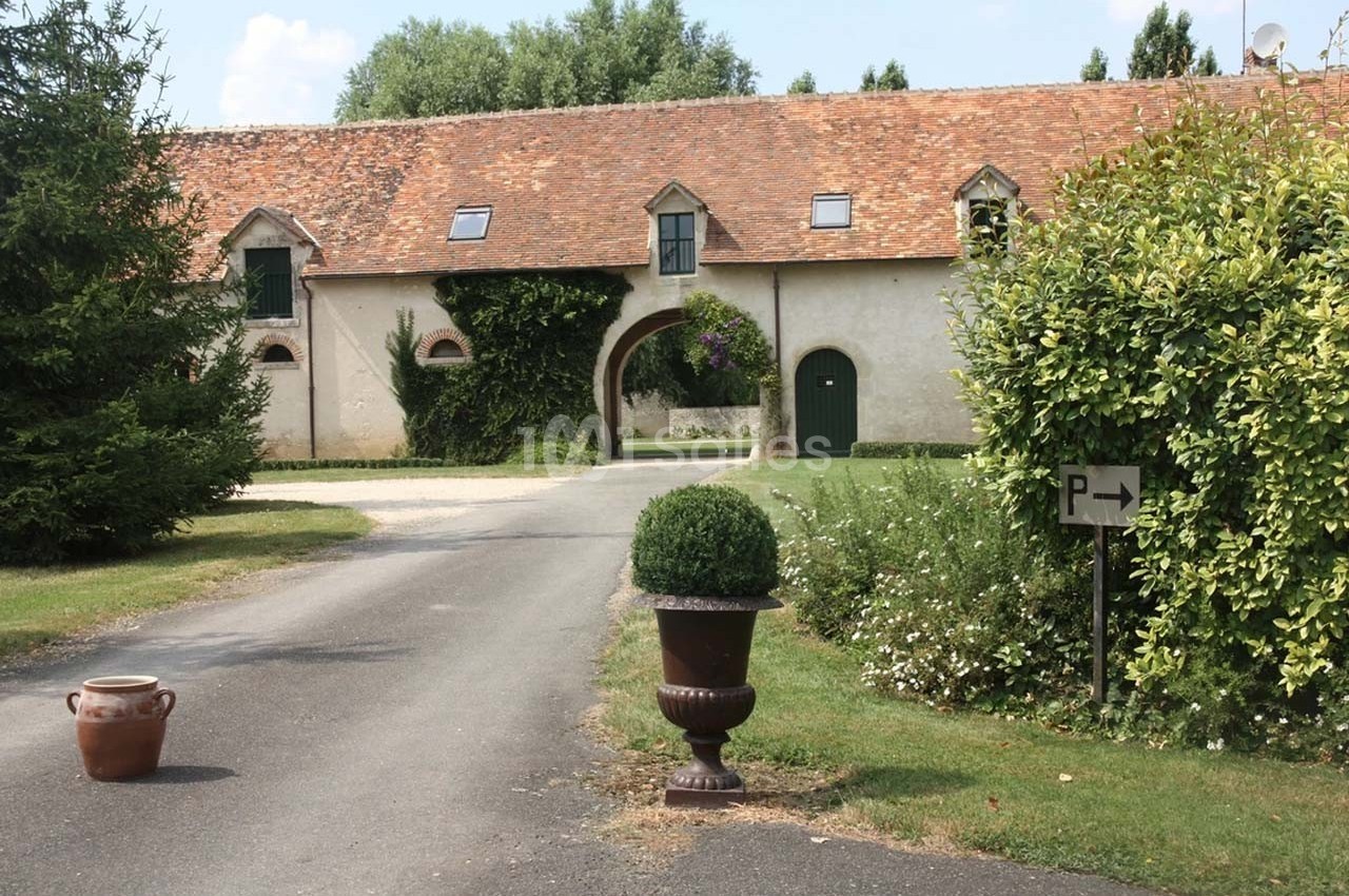 Allée menant à un bâtiment en pierre avec toit en tuiles, entouré de végétation et d'un panneau indiquant un parking.
