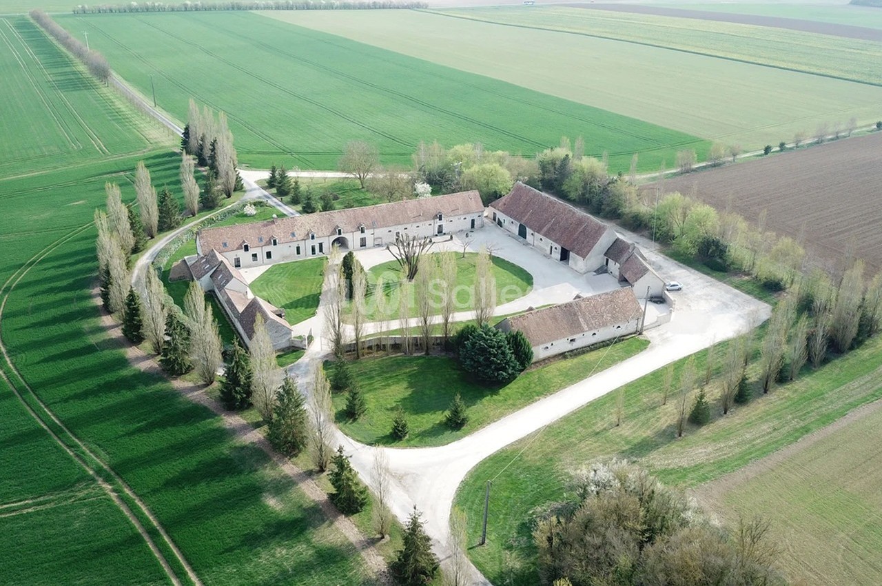 Vue aérienne d'une grande ferme en U entourée de champs verdoyants et de chemins bordés d'arbres.