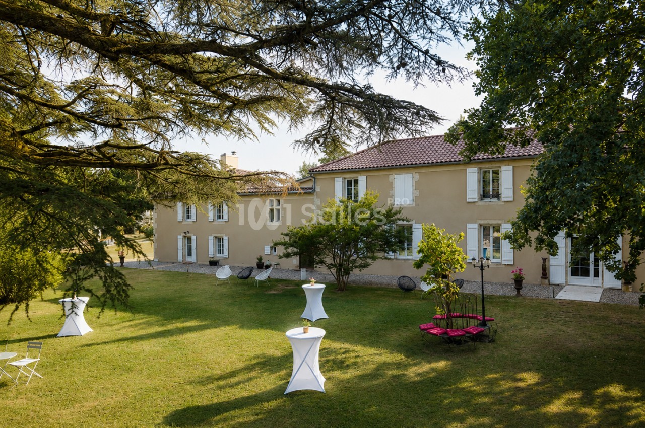 Grande maison en pierre avec jardin arboré, tables hautes blanches et mobilier de détente sur la pelouse.