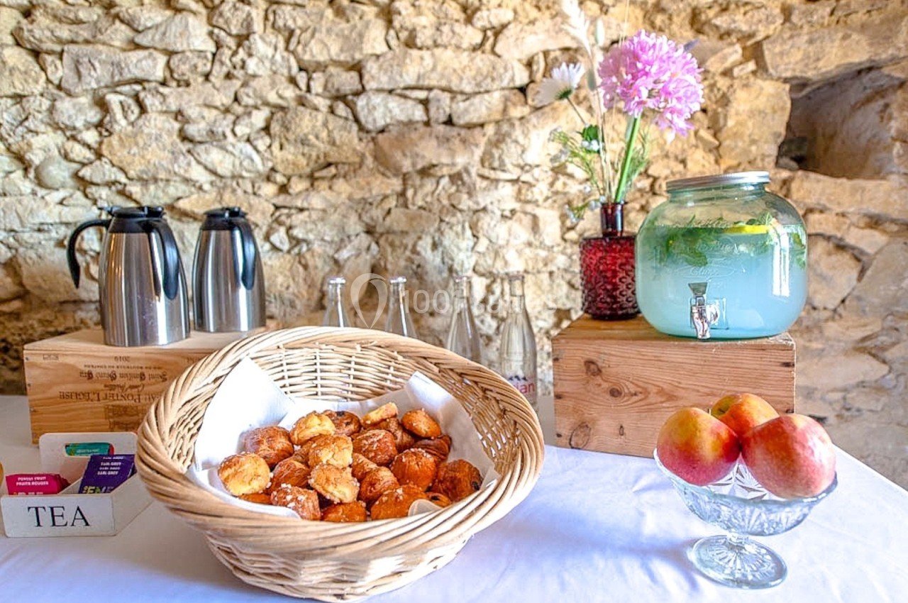 Table avec des viennoiseries, carafes, pommes et boissons, disposée devant un mur en pierre.