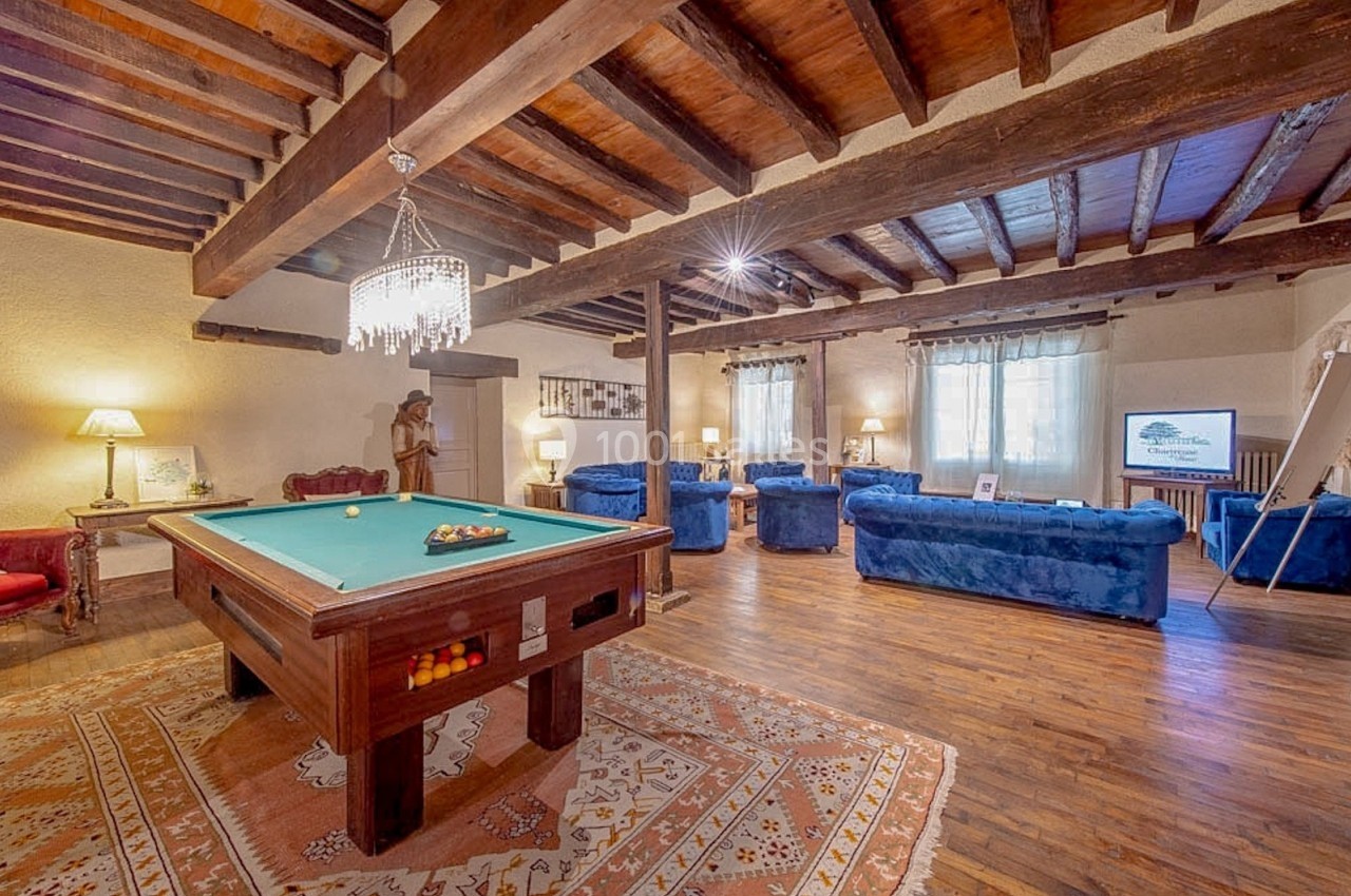 Salle de séjour spacieuse avec billard, canapés bleus, poutres apparentes et lumière naturelle.
