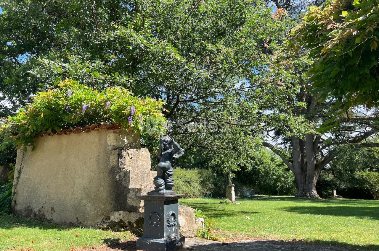 Statue en métal noir devant un mur ancien couvert de végétation, dans un jardin verdoyant sous un grand arbre.