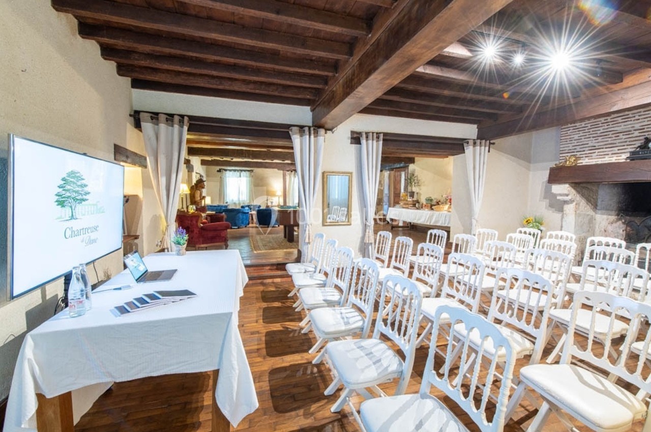 Salle de réunion avec chaises blanches alignées, table équipée d'un écran et plafond en poutres apparentes.