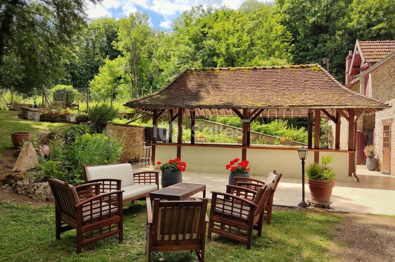 Salon de jardin en bois avec coussins blancs, installé sur une pelouse devant une terrasse couverte, entouré de verdure.