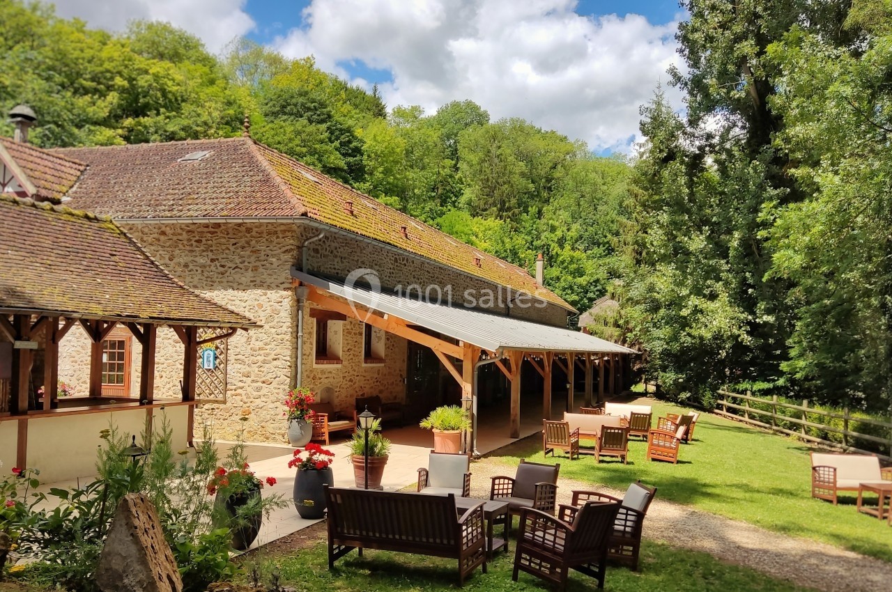 Bâtiment en pierre avec terrasse couverte, entouré de verdure et de mobilier de jardin sous un ciel partiellement nuageux.