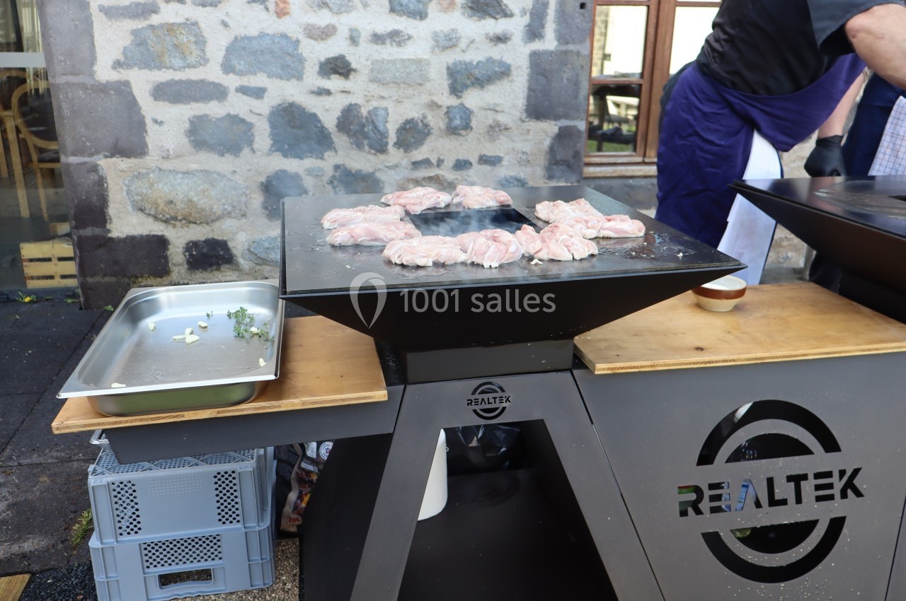 Cuisson de morceaux de viande sur une plancha en extérieur, avec un mur en pierre en arrière-plan.